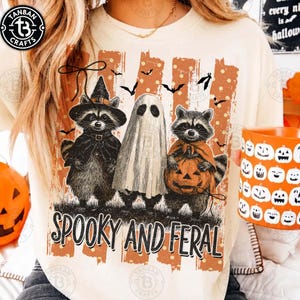 May include: A cream-colored t-shirt featuring a graphic of two raccoons and a ghost. One raccoon wears a witch hat, the other holds a pumpkin. The design includes orange vertical stripes, polka dots, bats, and the text "SPOOKY AND FERAL" in black.