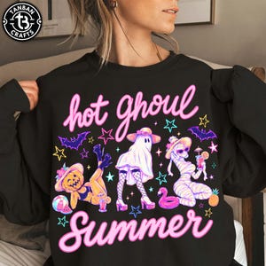 Op de afbeelding: Zwarte sweatshirt met de tekst "hot ghoul Summer" in roze script. De afbeelding toont cartoon ghouls, waaronder een pompoen, een spook en een mummie, met sterren en vleermuizen. De sweatshirt heeft een ronde hals en lange mouwen.