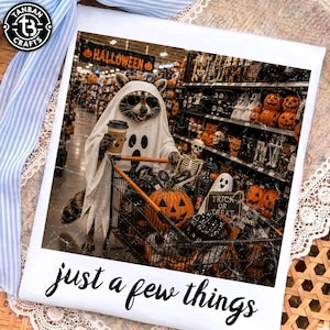 May include: A white t-shirt featuring a graphic of a raccoon wearing a ghost costume and sunglasses, holding a coffee cup labeled Boo Brew. The raccoon pushes a shopping cart filled with pumpkins and skeletons. Text reads just a few things.