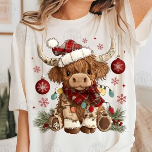 May include: A cream-colored t-shirt featuring a festive Highland cow illustration. The cow wears a Santa hat, a plaid scarf, and is adorned with Christmas lights and ornaments. Red snowflakes and ornaments complete the holiday design.