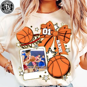 May include: Cream-colored t-shirt featuring basketball-themed graphics. The design includes orange basketballs, a large orange bow with the number "01", and a space for a name. A framed photo of basketball players is also included.