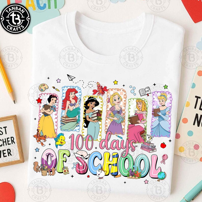 May include: White t-shirt featuring a colorful graphic celebrating "100 Days of School." The design includes illustrations of Disney princesses, books, and other school-related elements. The text "100 days of school" is prominently displayed in a playful font.
