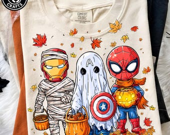 May include: A cream-coloured t-shirt featuring a graphic of three characters dressed in Halloween costumes: Iron Man as a mummy, Captain America as a ghost, and Spider-Man holding a jack-o'-lantern, all surrounded by falling autumn leaves.