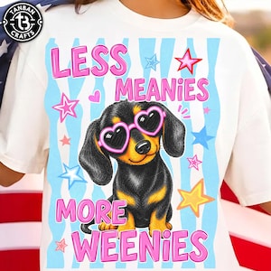 May include: White t-shirt with a graphic of a dachshund wearing heart-shaped sunglasses. The text reads "LESS MEANIES MORE WEENIES" in pink. The design includes stars and hearts on a blue and white striped background.