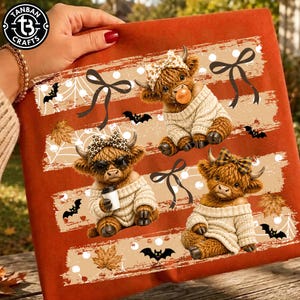 May include: An orange fabric item featuring a graphic print of three fluffy Highland cows wearing beige sweaters and patterned bows. The design includes black bats, white spiderwebs, and autumn leaves arranged in horizontal stripes across the surface.