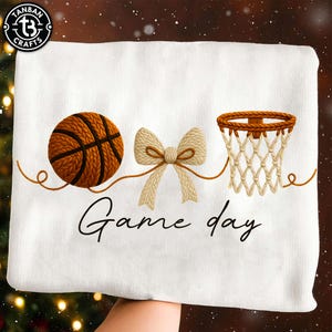 May include: White towel with a basketball, bow, and basketball hoop design. The words "Game day" are written in a script font below the images. The basketball is brown with black lines, and the bow is beige.
