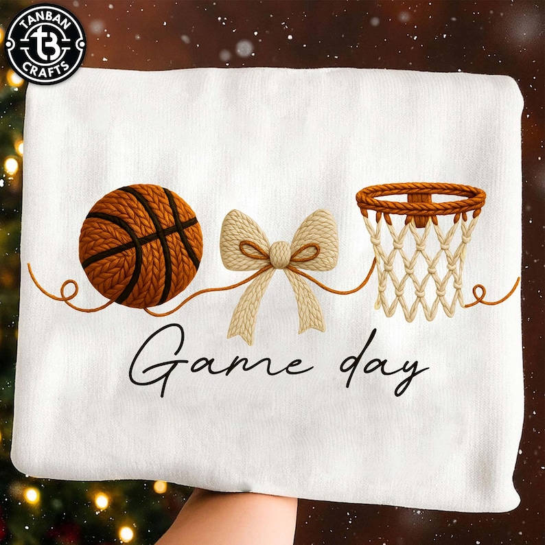 May include: White towel with a basketball, bow, and basketball hoop design. The words "Game day" are written in a script font below the images. The basketball is brown with black lines, and the bow is beige.