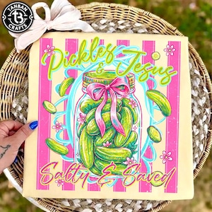 Op de afbeelding: Vierkante canvas print met een pot augurken met een roze strik, omringd door zwevende augurkenschijfjes en bloemen. De tekst luidt "Pickles & Jesus Salty & Saved" in een grillig lettertype, tegen een roze en crème gestreepte achtergrond.