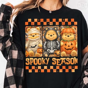 May include: A black t-shirt featuring a graphic of three cartoon bears dressed for Halloween as a mummy, a skeleton, and a pumpkin. The design includes orange and black checkered borders and the text SPOOKY SEASON in orange block letters.