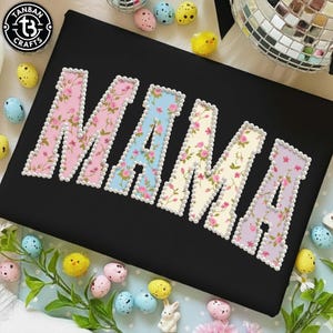 May include: Black fabric with the word "MAMA" in large, floral-patterned letters. Each letter is outlined with white pearls. The letters are in pink, blue, cream, and lavender floral patterns.