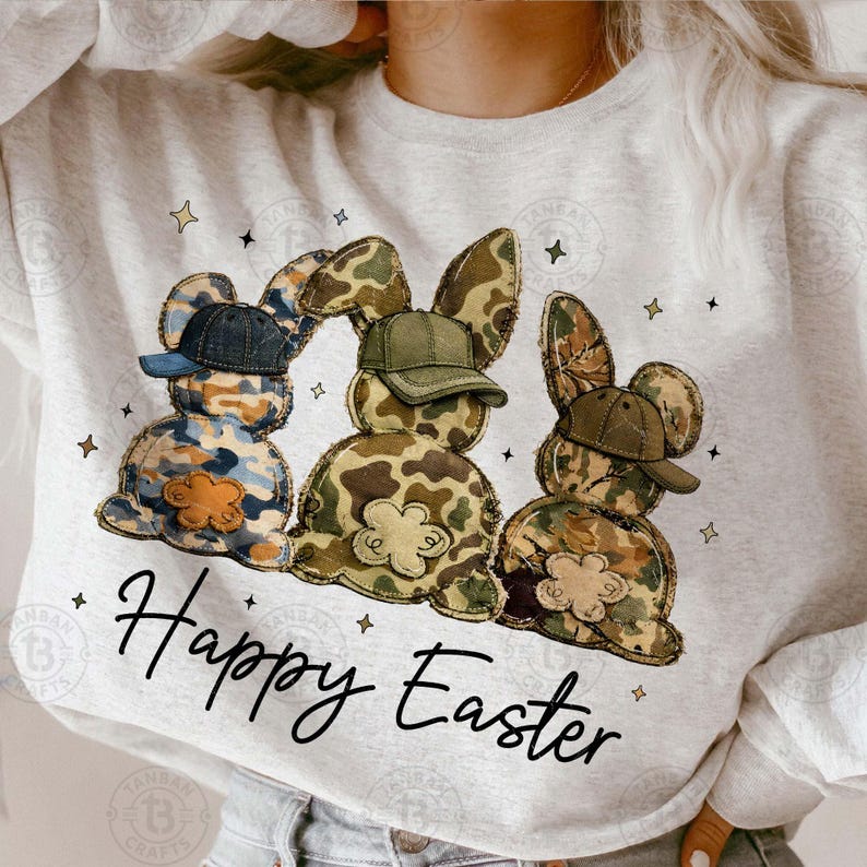 Op de afbeelding: Een lichtgrijs sweatshirt met drie konijnenfiguren met camouflagepatronen en baseballpetten. De woorden "Happy Easter" staan in een scriptletter onder de konijnen. Kleine stervormen zijn rond het ontwerp verspreid.