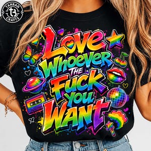Op de afbeelding: Een zwart t-shirt met een levendige, meerkleurige afbeelding met de tekst "LOVE WHOEVER THE FUCK YOU WANT" in een graffiti-stijl lettertype. Het ontwerp bevat regenboogkleurige elementen zoals een vlinder, planeet, cassettebandje, kersen, lippen, discobal en sterren.