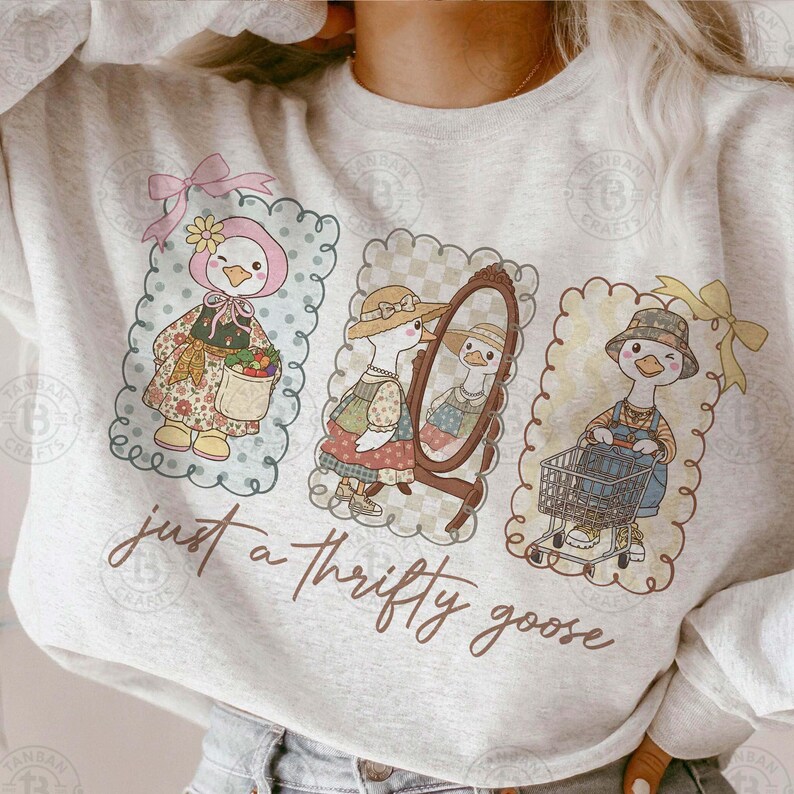 Puede incluir: Una sudadera gris claro con tres ilustraciones enmarcadas de un ganso. El ganso se representa con varios atuendos, incluyendo un gorro, un vestido y un sombrero, con el texto "just a thrifty goose" en escritura marr&oacute;n.