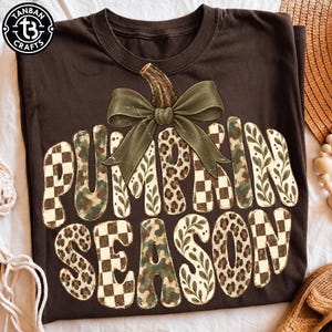 May include: A dark brown t-shirt featuring a large graphic of a pumpkin with a green bow. The text PUMPKIN SEASON is printed in large letters filled with various patterns including leopard print, camouflage, checkered, and leaf designs.