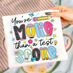 K&ouml;nnte beinhalten: Wei&szlig;es T-Shirt mit dem Text "You're MORE than a test SCORE" in farbenfrohen, verspielten Buchstaben. Das Design beinhaltet Bleistifte, Sterne, Blumen und einen Apfel, was ein schulisches Thema ergibt.