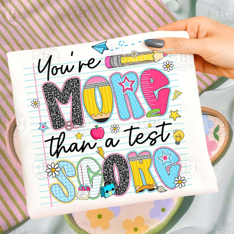 K&ouml;nnte beinhalten: Wei&szlig;es T-Shirt mit dem Text "You're MORE than a test SCORE" in farbenfrohen, verspielten Buchstaben. Das Design beinhaltet Bleistifte, Sterne, Blumen und einen Apfel, was ein schulisches Thema ergibt.