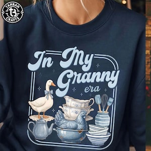 May include: Navy blue sweatshirt with the text "In My Granny era" in light blue. The design includes a goose with a bow, teacups, a chicken, and kitchen utensils. The Tanban Crafts logo is in the upper left corner.