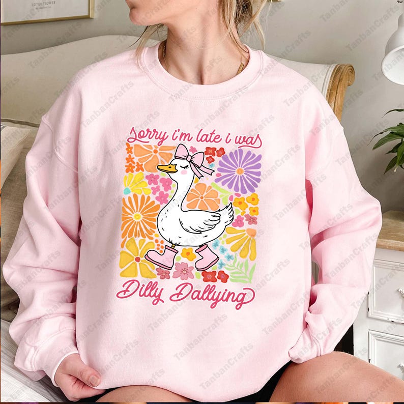 May include: Pink sweatshirt with a white duck wearing pink boots and a bow. The duck is surrounded by colorful flowers. The text on the sweatshirt reads "Sorry I'm late I was Dilly Dallying."