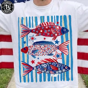May include: White t-shirt featuring three decorative fish with patriotic designs. The fish have red, white, and blue patterns, including stars, stripes, and floral elements. Vertical blue stripes with gold accents frame the fish.