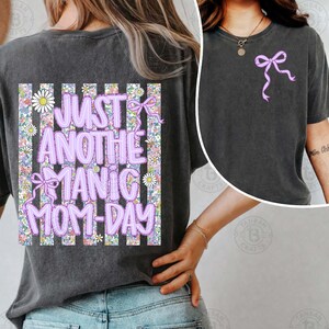 May include: Dark gray t-shirt with a lilac bow graphic on the front and the text "JUST ANOTHER MANIC MOM-DAY" on the back. The back design features a floral pattern with daisies and a lilac bow.
