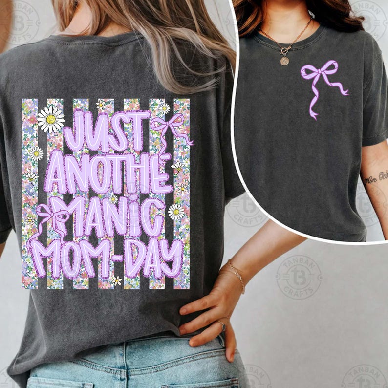 May include: Dark gray t-shirt with a lilac bow graphic on the front and the text "JUST ANOTHER MANIC MOM-DAY" on the back. The back design features a floral pattern with daisies and a lilac bow.