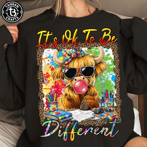 May include: Black sweatshirt with a colorful graphic of a Highland cow wearing sunglasses and blowing a bubble. The text "It's Ok To Be Different" is displayed in a rainbow font. The cow is surrounded by colorful paint splatters.