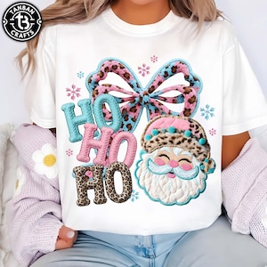 May include: White t-shirt with a festive Christmas design. The design features the words "HO HO HO" in blue, pink, and leopard print, a bow, snowflakes, and a Santa Claus face wearing a leopard print hat.