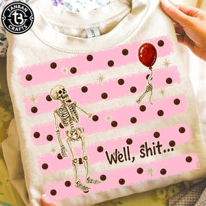 May include: A cream-colored sweatshirt featuring a graphic print of a skeleton holding a red balloon. The design includes horizontal pink stripes with dark brown polka dots and small gold star accents. The text "Well, shit..." is printed in brown.