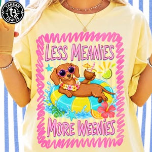 May include: Yellow t-shirt with a graphic of a dachshund wearing sunglasses and a flower lei, floating on an inner tube. The text reads "LESS MEANIES MORE WEENIES" in pink. A coconut drink and tropical flowers are also featured.
