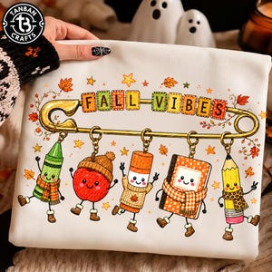 以下が含まれることがあります： 大きな金の安全ピンと「FALL VIBES」の文字が描かれた白いスウェットシャツ。安全ピンには、緑のクレヨン、赤いリンゴ、スティックのり、ノート、黄色い鉛筆という5つのキャラクターがぶら下がっています。