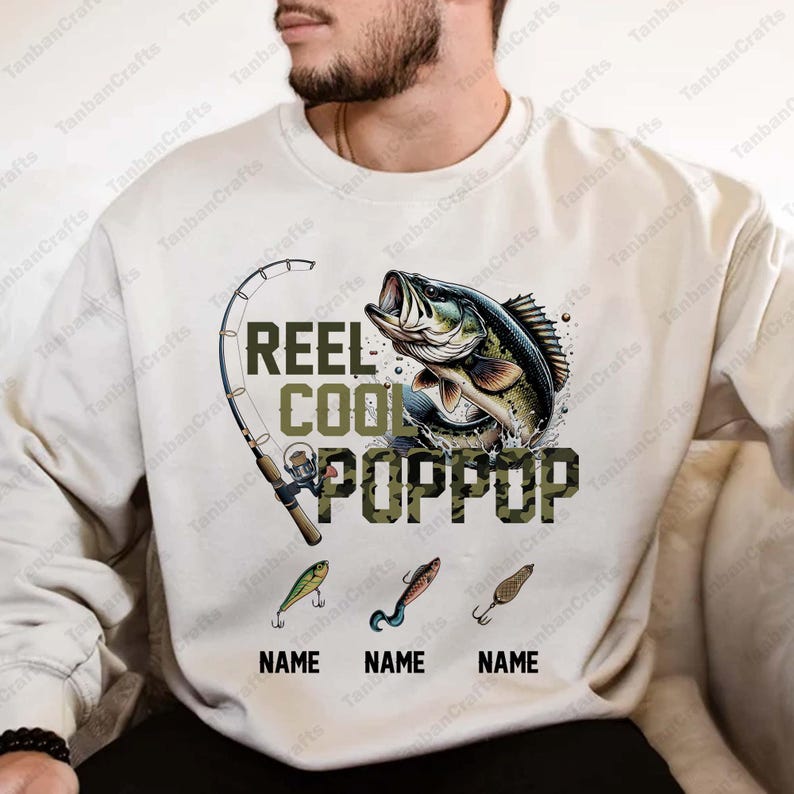 Pu&ograve; includere: Felpa color crema con un tema di pesca. Il design include una canna da pesca, un pesce che salta e le parole "REEL COOL POPPOP" in un font mimetico. Sotto ci sono tre esche da pesca e spazi per i nomi.