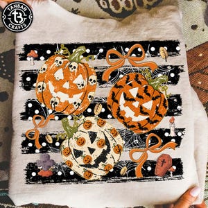 May include: A beige sweatshirt featuring a graphic print of three jack-o'-lanterns, orange ribbons, spiderwebs, a tombstone, and a coffin. The pumpkins are decorated with skull, bat, and small pumpkin patterns against black horizontal stripes.