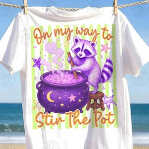May include: A white t-shirt featuring a graphic of a purple raccoon standing on a small wooden stool, stirring a purple cauldron with a wooden spoon. The text "On my way to Stir The Pot" is printed in orange, with light green vertical stripes.