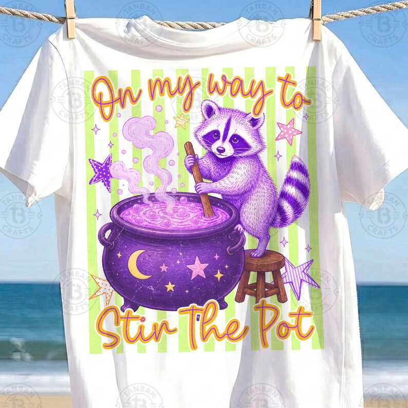 May include: A white t-shirt featuring a graphic of a purple raccoon standing on a small wooden stool, stirring a purple cauldron with a wooden spoon. The text "On my way to Stir The Pot" is printed in orange, with light green vertical stripes.
