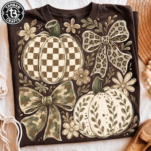 May include: A dark brown t-shirt featuring a graphic print of two pumpkins, two bows, and several flowers. One pumpkin has a brown and white checkered pattern, while the other has a white surface with green leaf accents. One bow has a leopard print, and the other has a camouflage pattern. The logo TANBAN CRAFTS is in the top left corner.