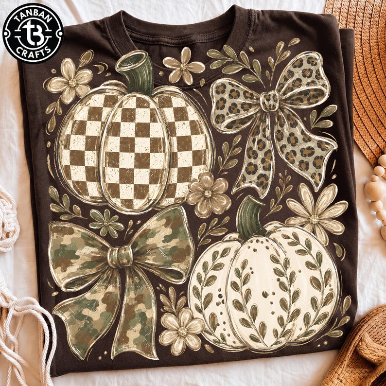 May include: A dark brown t-shirt featuring a graphic print of two pumpkins, two bows, and several flowers. One pumpkin has a brown and white checkered pattern, while the other has a white surface with green leaf accents. One bow has a leopard print, and the other has a camouflage pattern. The logo TANBAN CRAFTS is in the top left corner.