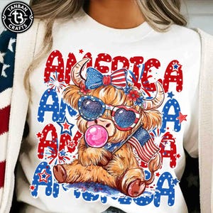 以下が含まれることがあります： サングラス、リボン、アメリカ国旗を身に着けた漫画のハイランド牛が描かれた白いTシャツ。牛はガムバブルを吹いています。「AMERICA」の文字が赤と青でプリントされています。