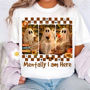 May include: White t-shirt featuring a graphic with three ghost figures in different settings, holding drinks. The text "Mentally I am Here" is below the images. The design includes a sign that says "Beach, Lake, Sunshine".