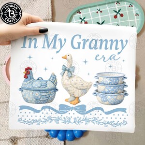 May include: A white print with the text "In My Granny era" and illustrations of a blue chicken-shaped container, a goose with a blue bow, and three blue floral bowls. The print has a blue ribbon and floral border.