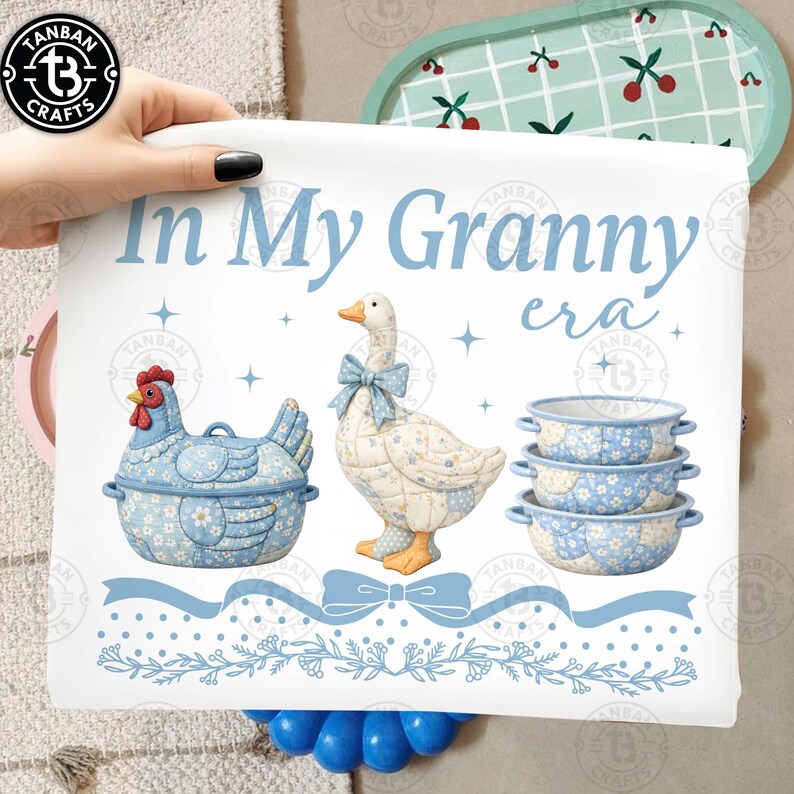 May include: A white print with the text "In My Granny era" and illustrations of a blue chicken-shaped container, a goose with a blue bow, and three blue floral bowls. The print has a blue ribbon and floral border.