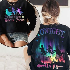 Puede incluir: Sudadera negra con el texto "IT'S JUST A BUNCH OF Hocus Pocus" y un gr&aacute;fico de tres brujas volando en escobas. La parte trasera de la sudadera dice "TONIGHT We fly". El dise&ntilde;o presenta un degradado colorido.