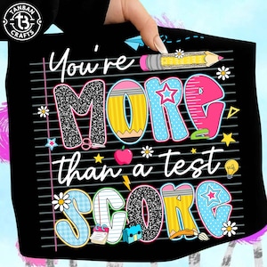 K&ouml;nnte beinhalten: Schwarzes T-Shirt mit dem Text "You're MORE than a test SCORE" in farbenfrohen, verspielten Buchstaben. Das Design umfasst Bleistifte, Sterne, Blumen und einen Apfel und schafft eine schulische Grafik.