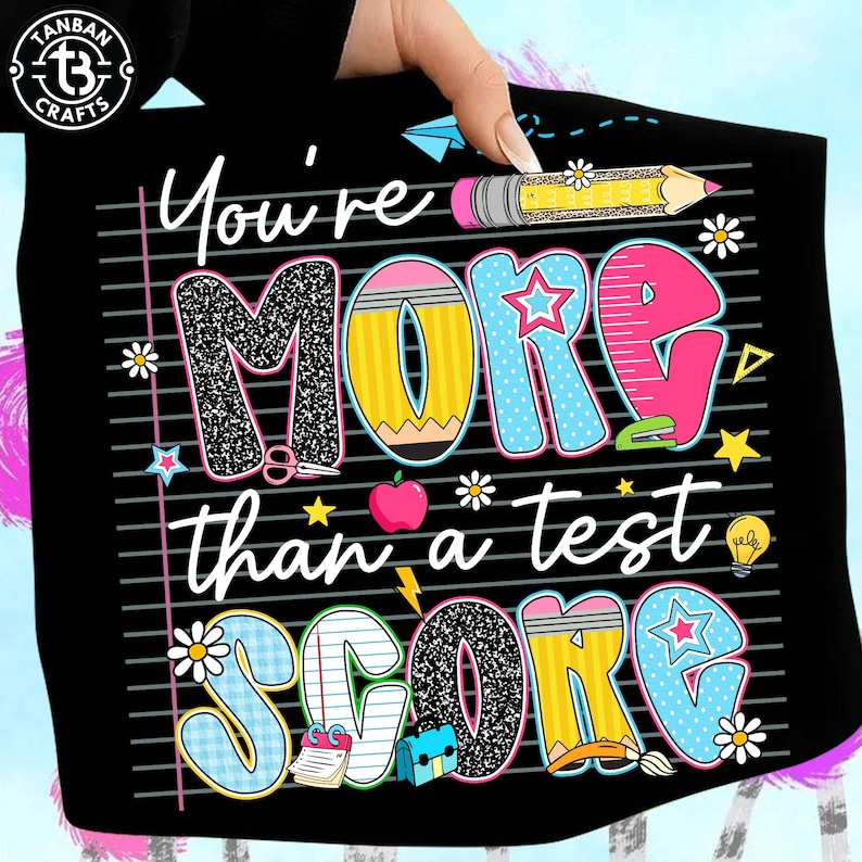 K&ouml;nnte beinhalten: Schwarzes T-Shirt mit dem Text "You're MORE than a test SCORE" in farbenfrohen, verspielten Buchstaben. Das Design umfasst Bleistifte, Sterne, Blumen und einen Apfel und schafft eine schulische Grafik.