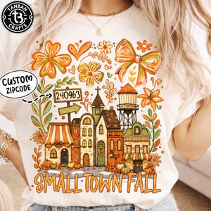 May include: A cream-colored t-shirt featuring a graphic print of a small town scene with buildings, a water tower, pumpkins, and flowers in orange, green, and brown tones. The text reads 240963 on a sign and SMALL TOWN FALL at the bottom.