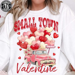 Puede incluir: Sudadera blanca con un diseño de San Valentín. Presenta una camioneta roja que transporta un ramo de flores y un grupo de globos en forma de corazón en tonos rojos, rosas y crema. Las palabras "Small Town Valentine" están impresas en rojo.