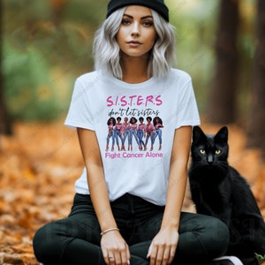 May include: White t-shirt with the text "S.I.S.T.E.R.S don't let sisters Fight Cancer Alone" in pink. The shirt features an illustration of six women in jeans and pink tops. A black cat sits beside the person wearing the shirt.