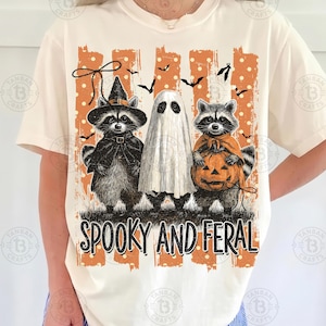 May include: A cream-colored t-shirt featuring a graphic of two raccoons and a ghost dressed for Halloween. One raccoon wears a witch hat, the other holds a jack-o'-lantern. The design includes orange stripes, polka dots, bats, and the text SPOOKY AND FERAL.