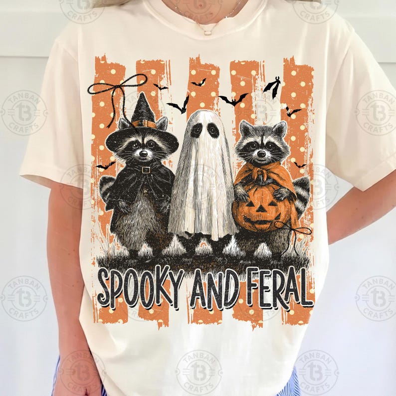 May include: A cream-colored t-shirt featuring a graphic of two raccoons and a ghost dressed for Halloween. One raccoon wears a witch hat, the other holds a jack-o'-lantern. The design includes orange stripes, polka dots, bats, and the text SPOOKY AND FERAL.