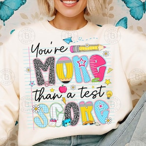 K&ouml;nnte beinhalten: Cremefarbenes Sweatshirt mit einem farbenfrohen Grafikdesign mit der Aufschrift "You're MORE than a test SCORE". Das Design umfasst einen Bleistift, Sterne, Blumen und einen Apfel. Ein l&auml;ssiger, bequemer Kleidungsstil.