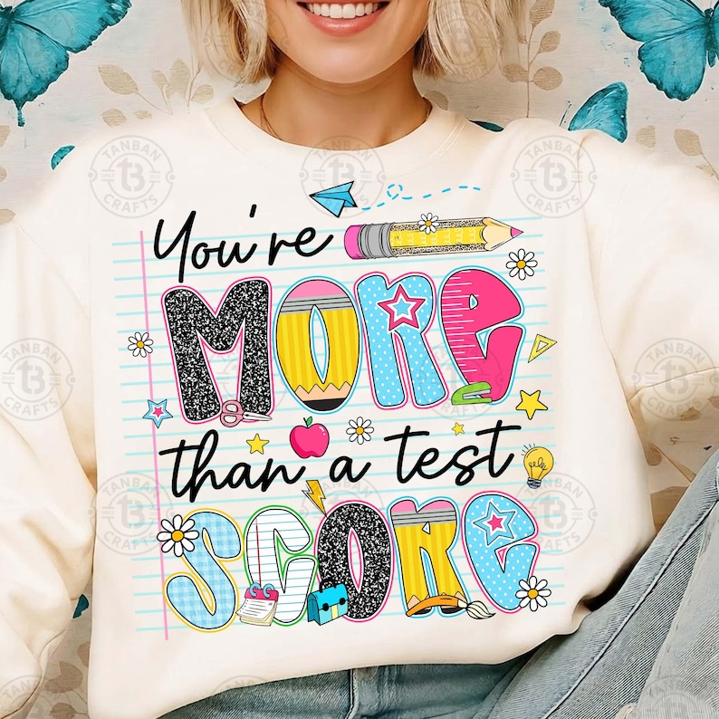 K&ouml;nnte beinhalten: Cremefarbenes Sweatshirt mit einem farbenfrohen Grafikdesign mit der Aufschrift "You're MORE than a test SCORE". Das Design umfasst einen Bleistift, Sterne, Blumen und einen Apfel. Ein l&auml;ssiger, bequemer Kleidungsstil.